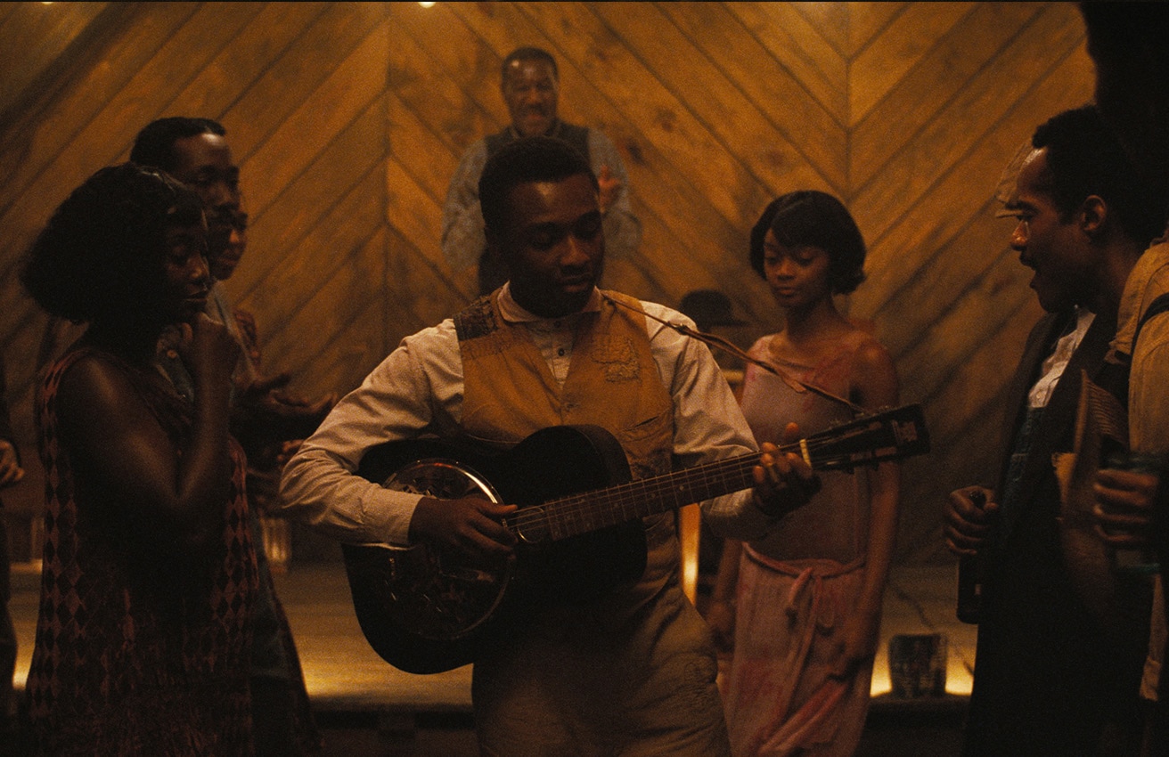 Sammie stands in the middle of a group of people in a dimly lit room and plays a resonator guitar. Delta Slim stands behind him on a stage and claps his hands.